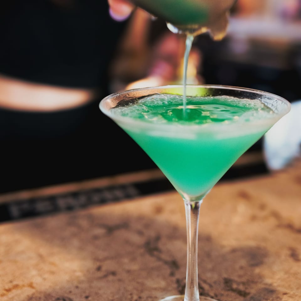 A bartender pours a vibrant green cocktail from a shaker into a martini glass at Nosh Kitchen & Cocktails.