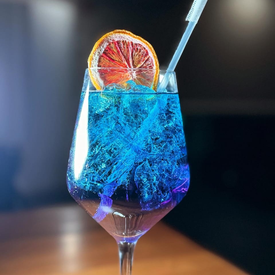 A vibrant blue cocktail with ice, garnished with a dried orange slice and a straw.