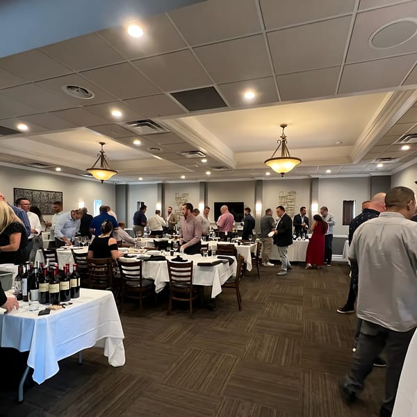 A large group of people mingling in a spacious dining room with set tables and wine bottles.