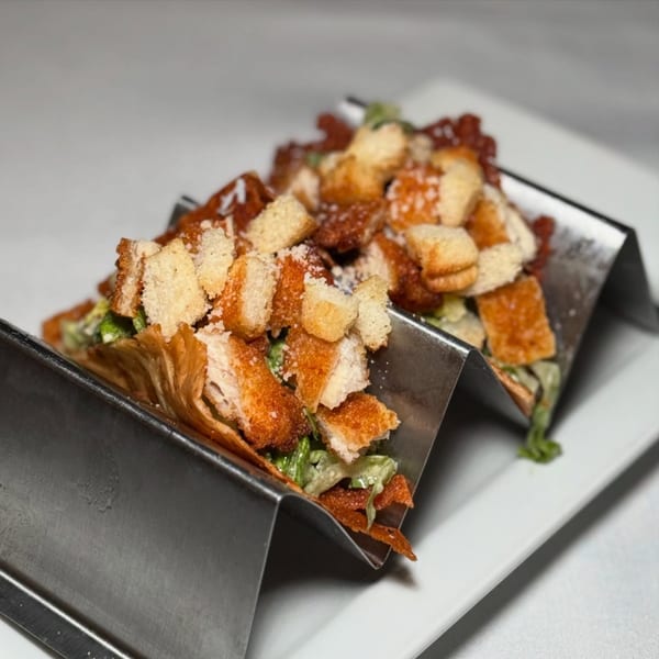 Three crispy chicken Caesar salad tacos in a metal holder on a white plate.