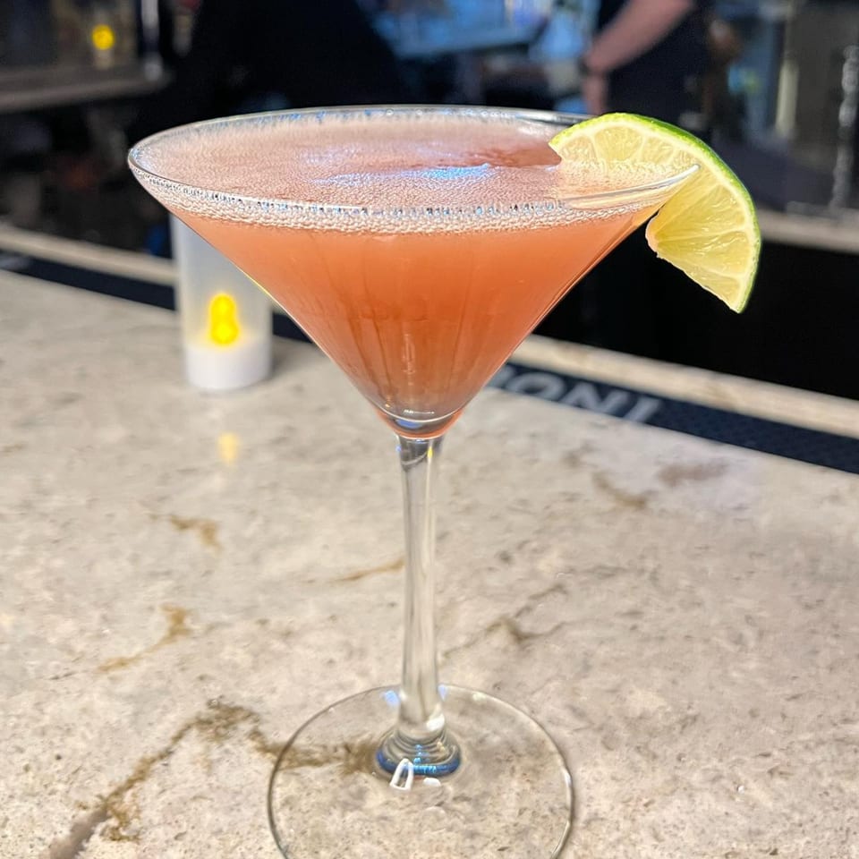 A pink cocktail in a martini glass with a lime wedge, on a bar counter at Nosh Kitchen & Cocktails.