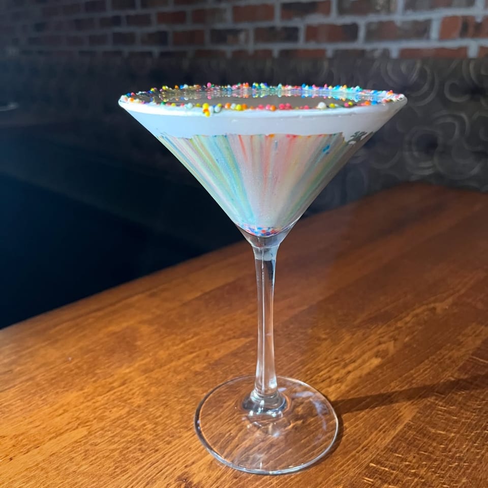 A colorful martini with rainbow sprinkles on the rim sits on a wooden table at Nosh Kitchen & Cocktails.