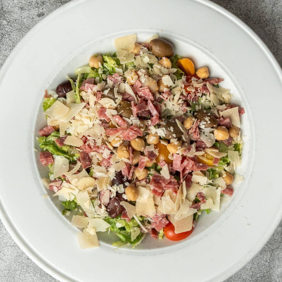 Overhead view of a Nosh Kitchen & Cocktails salad with lettuce, salami, chickpeas, olives, and parmesan.