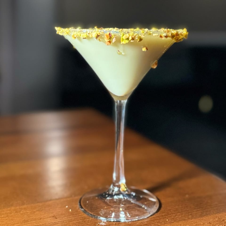 A Nosh Kitchen & Cocktails martini with a pistachio-crusted rim sits on a wooden table.
